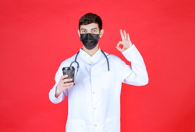 Médico con estetoscopio en máscara negra sosteniendo una taza de café para llevar negra y disfrutando del sabor.