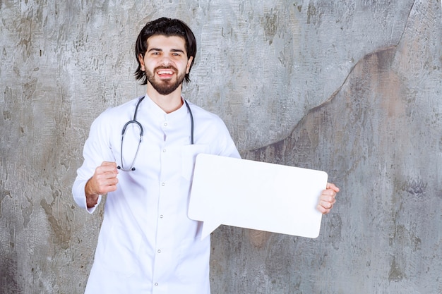 Médico con un estetoscopio sosteniendo un tablero de información en blanco de forma rectangular y mostrando su puño