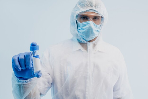 Médico europeu do sexo masculino mostrando a vacina COVID-19. Homem vestindo jaleco branco, óculos, máscara protetora e luva de látex. Conceito de combate ao coronavírus. Fundo cinza com luz turquesa. Sessão de estúdio