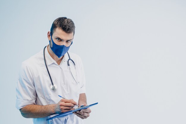 Médico europeu masculino na máscara protetora, escrevendo algo na área de transferência. Homem com estetoscópio usa jaleco branco. Pessoa com cara de cética. Fundo cinza com luz turquesa. Sessão de estúdio. Copie o espaço