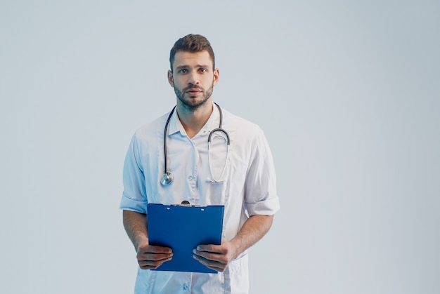 Médico europeu preocupado com estetoscópio e área de transferência. jovem barbudo homem vestindo jaleco branco. isolado em um fundo cinza com luz turquesa. sessão de estúdio. copie o espaço.