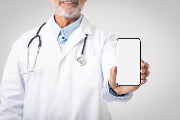 Médico europeu sênior sorridente em jaleco branco com estetoscópio mostra smartphone com tela em branco