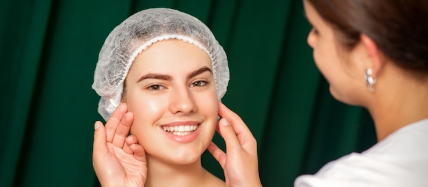 Médico examina el bello rostro de la joven mujer sonriente en la clínica de belleza.