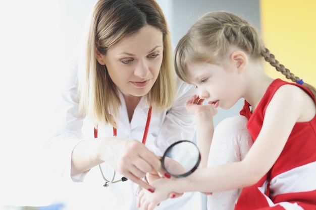 Médico examina la piel en la mano de la niña con lupa en la clínica