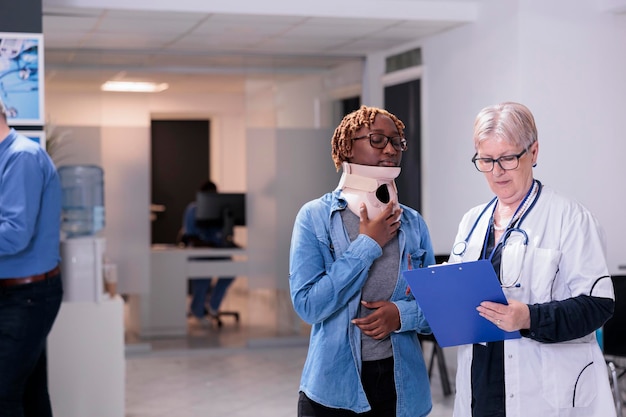 Médico examinando a una mujer con cuello, usando un aparato ortopédico de espuma cervical después de una lesión por accidente. Diversas personas que realizan consultas de chequeo con cita médica en el vestíbulo de la sala de espera.