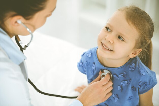 Foto médico examinando a un niño con estetoscopio. una paciente feliz y sonriente vestida con un vestido azul se encuentra en la inspección médica habitual. concepto de medicina.