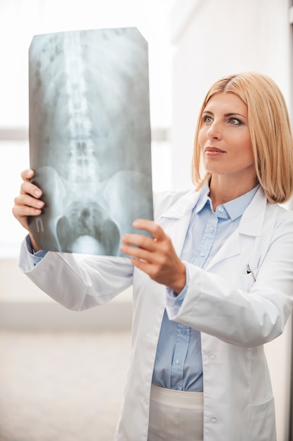 Médico examinando raio-x. Médica confiante de uniforme branco segurando uma imagem de raio-x e olhando para ela