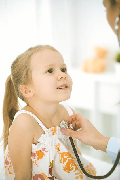 Médico examinando uma criança pelo estetoscópio. A paciente sorridente feliz vestida com um vestido de cor brilhante está na inspeção médica habitual.