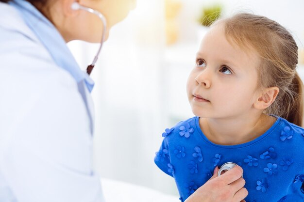 Médico examinando uma criança pelo estetoscópio na clínica ensolarada. Paciente menina sorridente feliz vestida de vestido azul está na inspeção médica habitual.