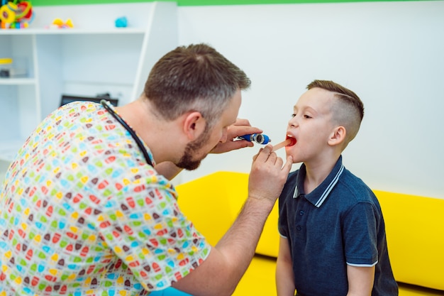 Médico experiente está olhando para a garganta do menino com instrumentos especiais na clínica. pediatra em uniforme médico examinando garganta criança.