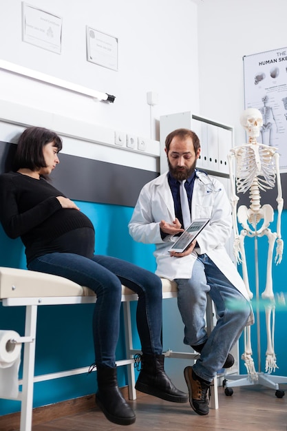 Médico explicando a varredura de osteopatia no tablet para paciente grávida. Mulher esperando filho, participando de consulta de check-up com clínico geral, olhando para o esqueleto humano na tela.