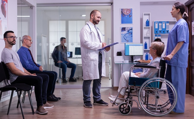 Médico falando com mulher com deficiência na sala de espera do hospital, lendo formulário de registro na prancheta