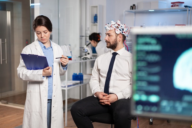 Médico falando sobre os sintomas da doença durante as investigações usando fone de ouvido de varredura de ondas cerebrais de alta tecnologia. Pesquisador analisando o estado de saúde do paciente homem, funções cerebrais, sistema nervoso.