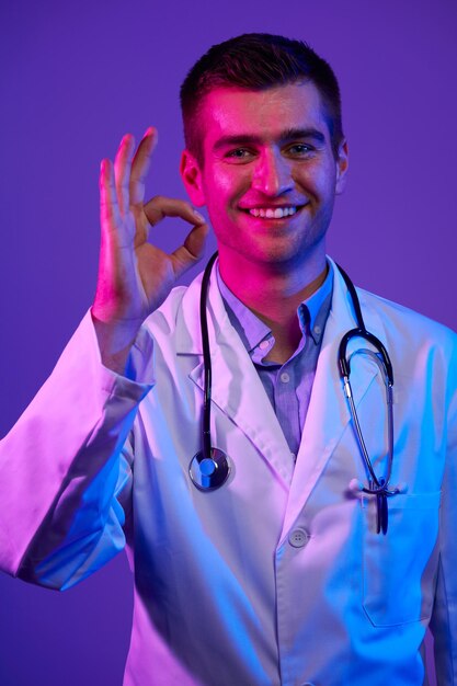 Foto médico fazendo sinal okey com os dedos, excelente símbolo luzes de néon fundo azul e rosa. pandemia do coronavírus