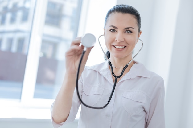 Médico feliz e satisfeito e confiante trabalhando no hospital, demonstrando o estetoscópio e expressando positividade