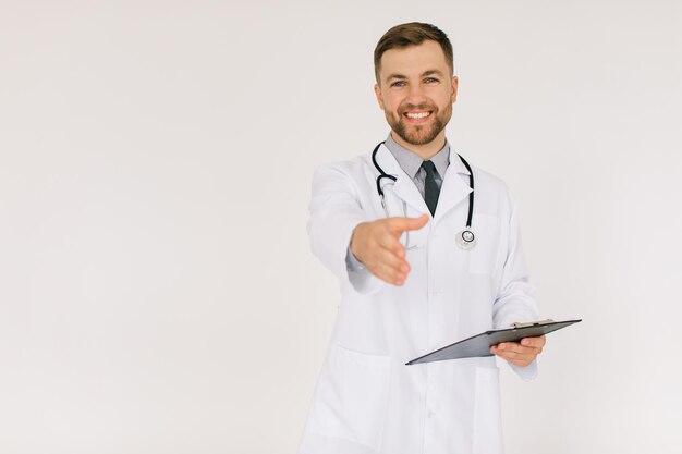 El médico feliz con un estetoscopio y una carpeta extiende una mano de saludos sobre un fondo blanco.