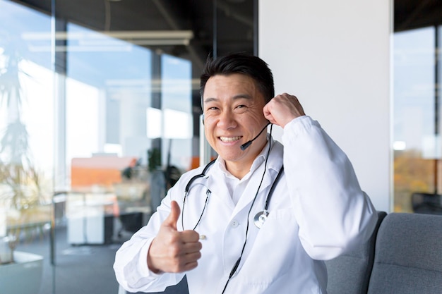 Médico feliz sorri e olha para a câmera Asiático anuncia boas notícias para o paciente usa uma chamada de vídeo e um fone de ouvido reunião on-line com colegas divertidos gesticulando com a visão da webcam das mãos