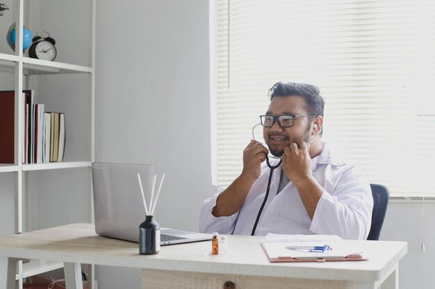 Médico feliz trabalhando com laptop em sua mesa enquanto segura seu estetoscópio