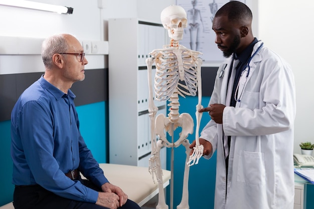 Médico fisioterapeuta mostrando esqueleto humano anatômico explicando dor nas costas para paciente idoso discutindo sintomas de doença durante fisioterapia médica no escritório do hospital. Tratamento da osteoporose