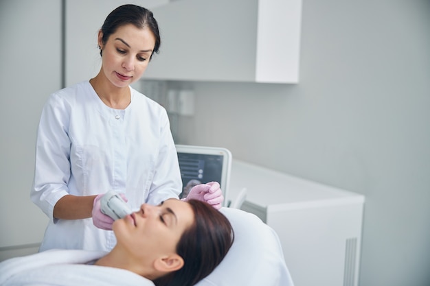 Médico focado olhando para o rosto de sua paciente durante o tratamento facial de ultrassom