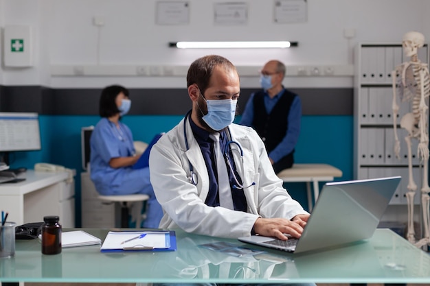 Médico general con bata blanca trabajando en una laptop en el consultorio médico. Especialista en atención médica que usa mascarilla facial y computadora para hacer un examen de experiencia profesional durante la pandemia.