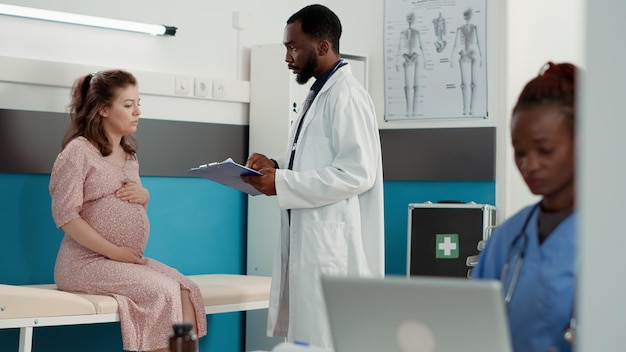 Médico general haciendo consulta de embarazo con futura madre, ayudando con el parto. Especialista en salud hablando con una mujer que espera un hijo en el consultorio médico, visita de control.