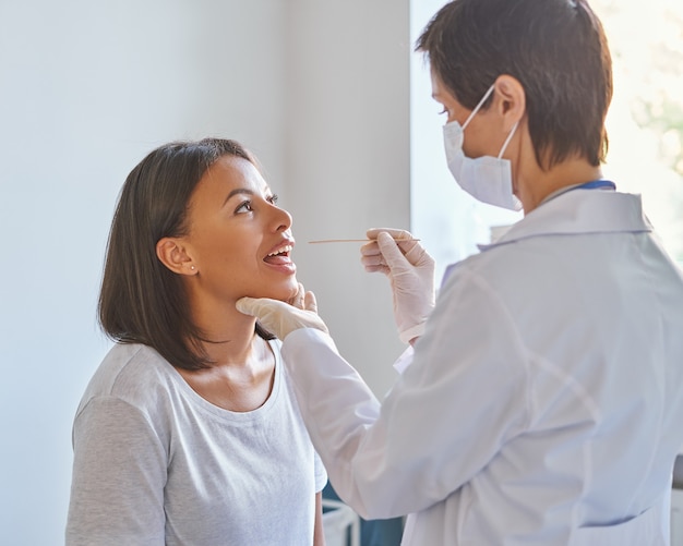 Foto médico general de mujer de mediana edad en máscara protectora facial examinando la garganta del paciente en la clínica