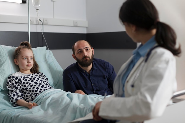 Médico general que consulta a un niño que se recupera mal sentado en la cama del paciente. Experto en pediatría hablando con el padre de una niña enferma hospitalizada descansando en la sala de un centro de salud infantil.