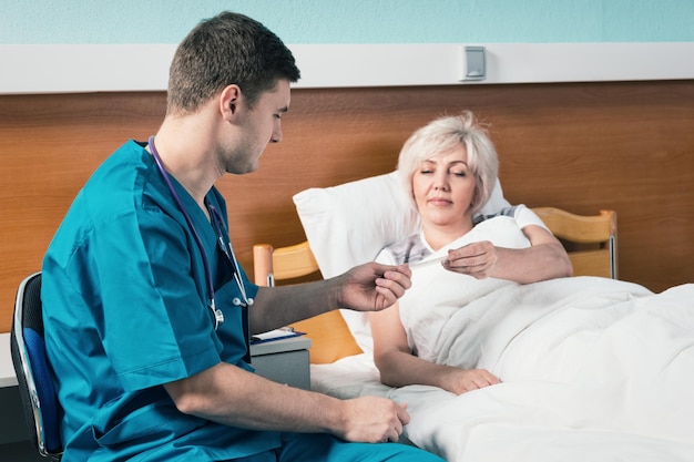 Médico guapo en uniforme con fonendoscopio en el cuello le da un termómetro a su paciente, que está acostado en la cama del hospital en la sala del hospital. Concepto de salud