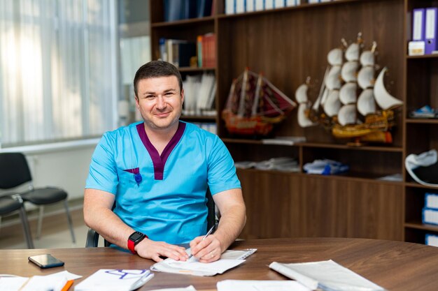 Médico hábil posando para la cámara mientras está sentado en la mesa en su gabinete Hombre guapo en uniforme médico Concepto de medicina