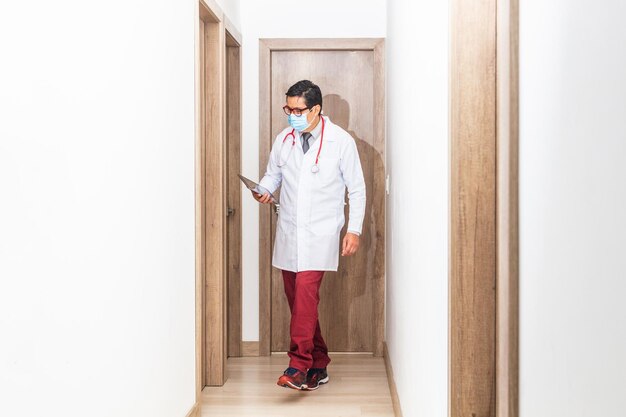 Médico hispano con una máscara de corbata blanca y un estetoscopio visitando a sus pacientes en un pasillo del hospital sosteniendo una tableta en sus manos Concepto de trabajadores de la salud durante una pandemia