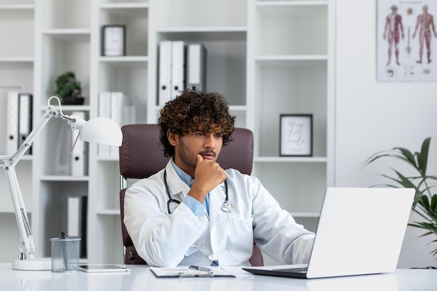 Un médico hispano serio y enfocado que trabaja dentro de un consultorio médico con un abrigo médico pensando en mirar