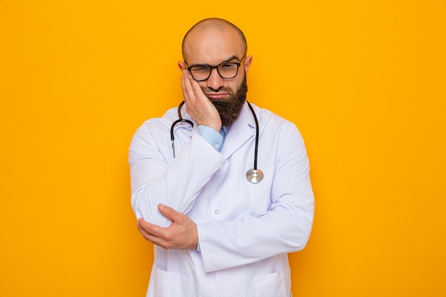 Médico hombre barbudo en bata blanca con estetoscopio alrededor del cuello con gafas con aspecto cansado y aburrido