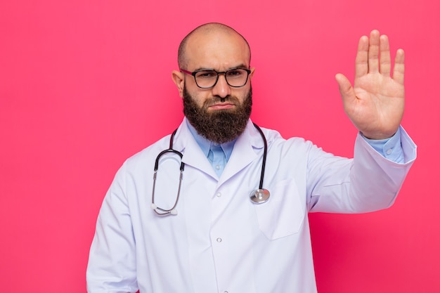 Médico hombre barbudo en bata blanca con estetoscopio alrededor del cuello con gafas mirando a la cámara con cara seria haciendo gesto de parada con la mano de pie sobre fondo rosa