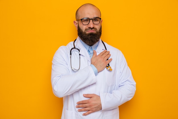 Médico hombre barbudo en bata blanca con estetoscopio alrededor del cuello con gafas mirando a la cámara sosteniendo la mano en el pecho sintiéndose agradecido de pie sobre fondo naranja