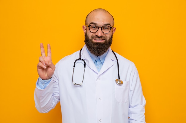 Médico hombre barbudo en bata blanca con estetoscopio alrededor del cuello con gafas mirando sonriendo alegremente mostrando v-sign