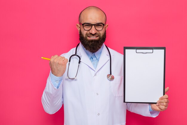 Médico homem barbudo com jaleco branco com estetoscópio em volta do pescoço usando óculos segurando a prancheta com páginas em branco feliz e animado punho cerrado sorrindo alegremente