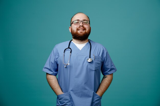 Médico homem barbudo de uniforme com estetoscópio no pescoço usando óculos feliz e satisfeito sorrindo amplamente de mãos dadas nos bolsos sobre fundo azul