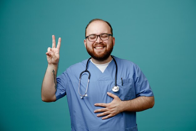 Médico homem barbudo de uniforme com estetoscópio no pescoço usando óculos feliz e satisfeito sorrindo amplamente mostrando vsign em pé sobre fundo azul