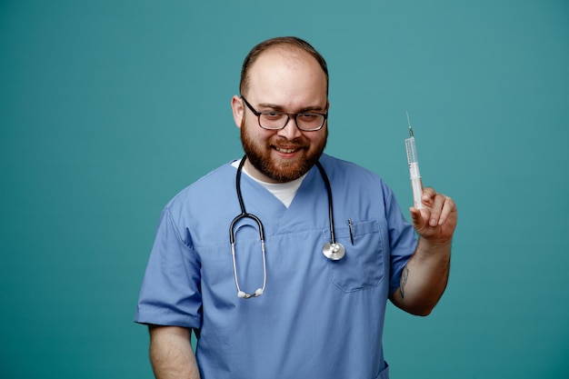 Médico homem barbudo de uniforme com estetoscópio no pescoço usando óculos segurando a seringa olhando para a câmera sorrindo maliciosamente em pé sobre fundo azul