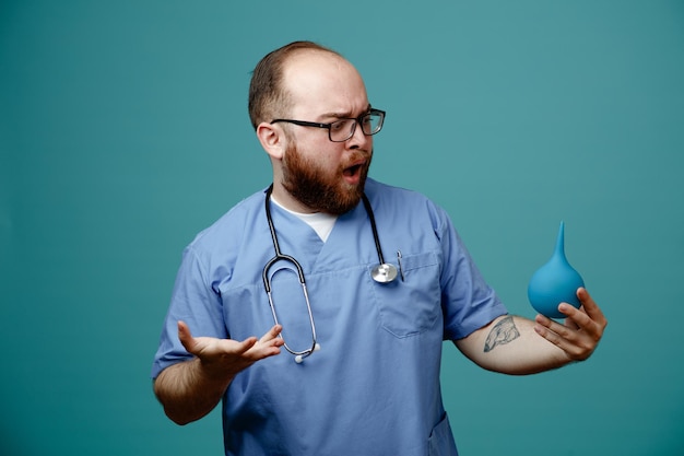 Médico homem barbudo de uniforme com estetoscópio no pescoço usando óculos segurando enema olhando confuso e desapontado em pé sobre fundo azul