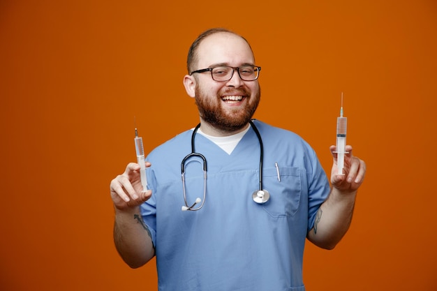 Médico homem barbudo de uniforme com estetoscópio no pescoço usando óculos segurando seringas sorrindo amplamente feliz e positivo em pé sobre fundo laranja