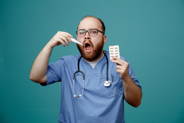 Médico homem barbudo de uniforme com estetoscópio no pescoço usando óculos segurando termômetro e pílulas olhando para a câmera com a boca aberta em pé sobre fundo azul