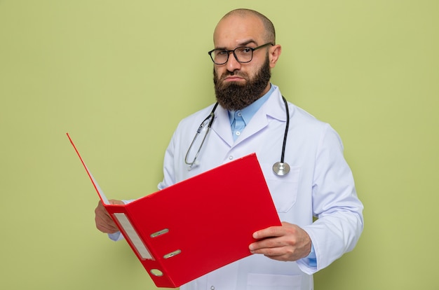 Médico homem barbudo vestindo jaleco branco com estetoscópio no pescoço usando óculos segurando uma pasta do escritório e olhando com uma cara séria