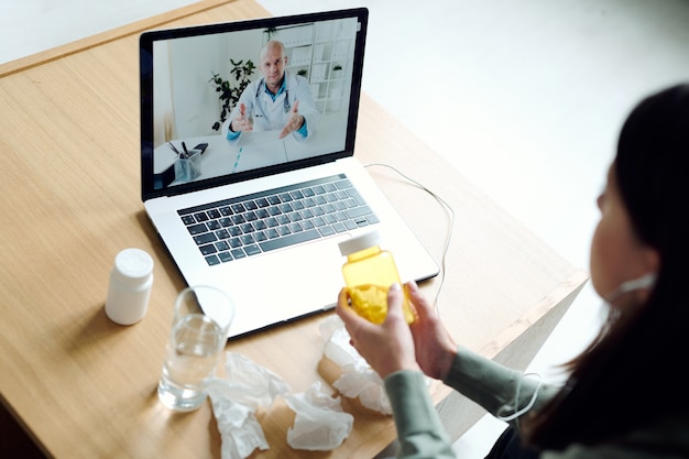 Foto médico homem maduro na tela do laptop dando recomendações médicas a uma jovem paciente doente do sexo feminino com um frasco de comprimidos e ouvindo seus conselhos