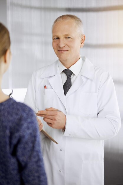 Foto médico idoso confiante consulta mulher-paciente na clínica ensolarada. serviço médico perfeito em medicina.