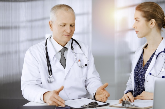 Médico idoso e sua colega discutindo a terapia da doença atual enquanto está sentado no local de trabalho na clínica ensolarada. Trabalho em equipe na medicina.