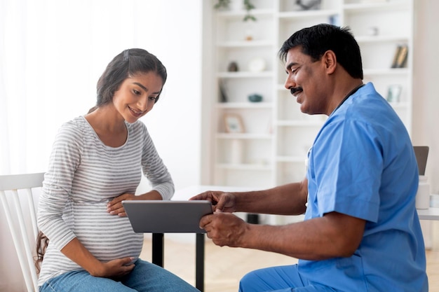 Médico indio maduro mostrando tableta digital a una mujer embarazada en la clínica