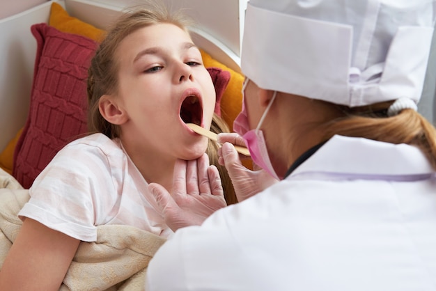 Médico infantil examinando a garganta da menina em casa com um abaixador de língua