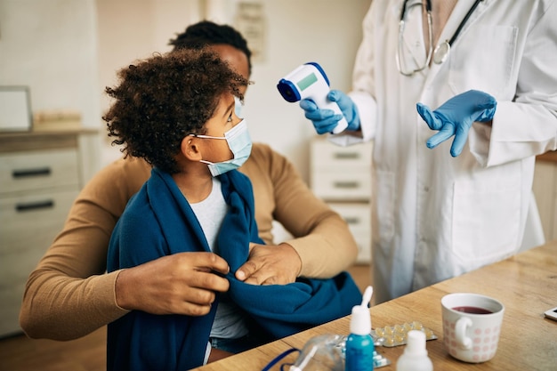 Médico irreconocible midiendo la temperatura de un niño pequeño con termómetro infrarrojo durante una visita a casa